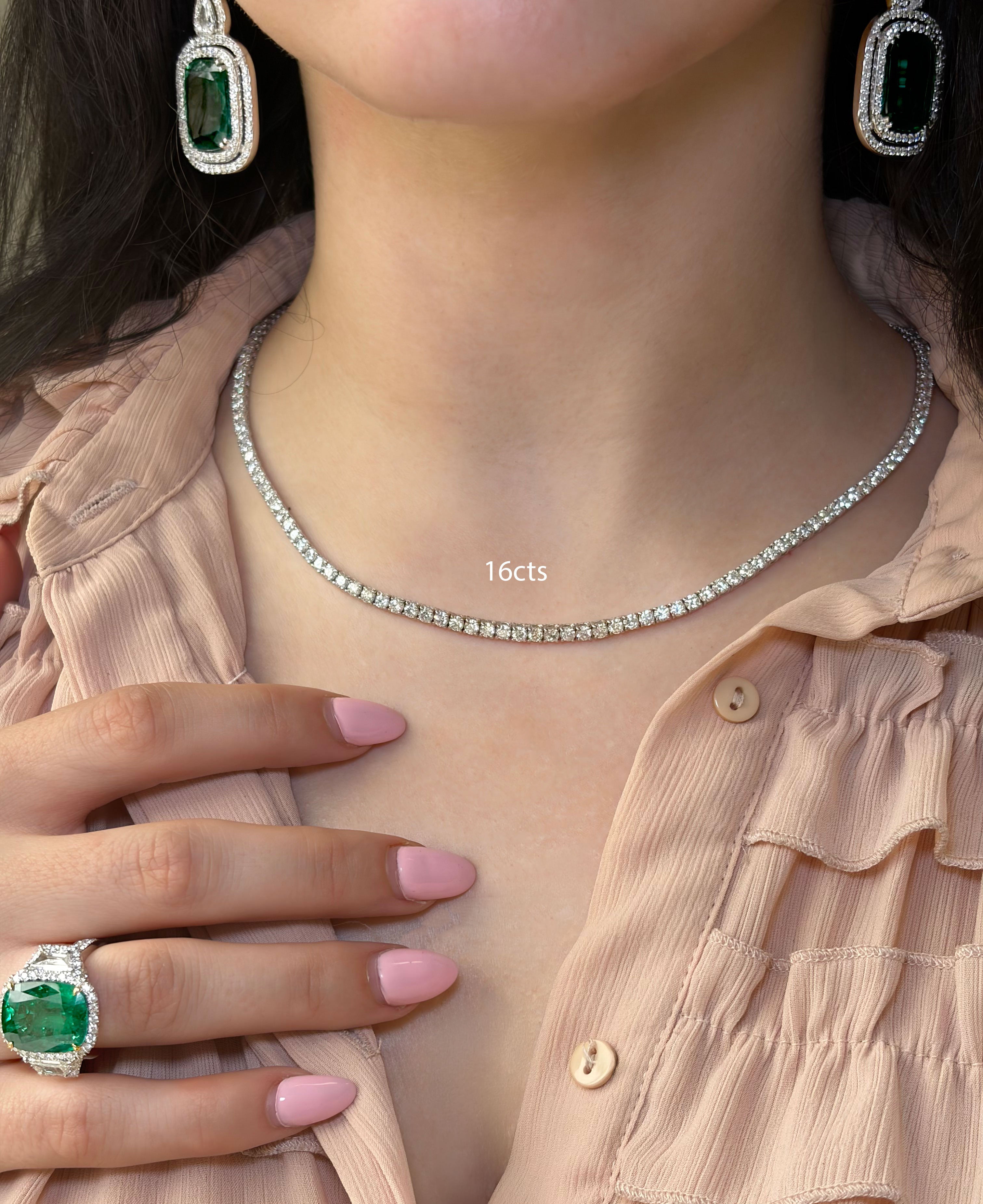 Close-up of a person wearing a diamond necklace, emerald earrings, and ring on a light pink background.