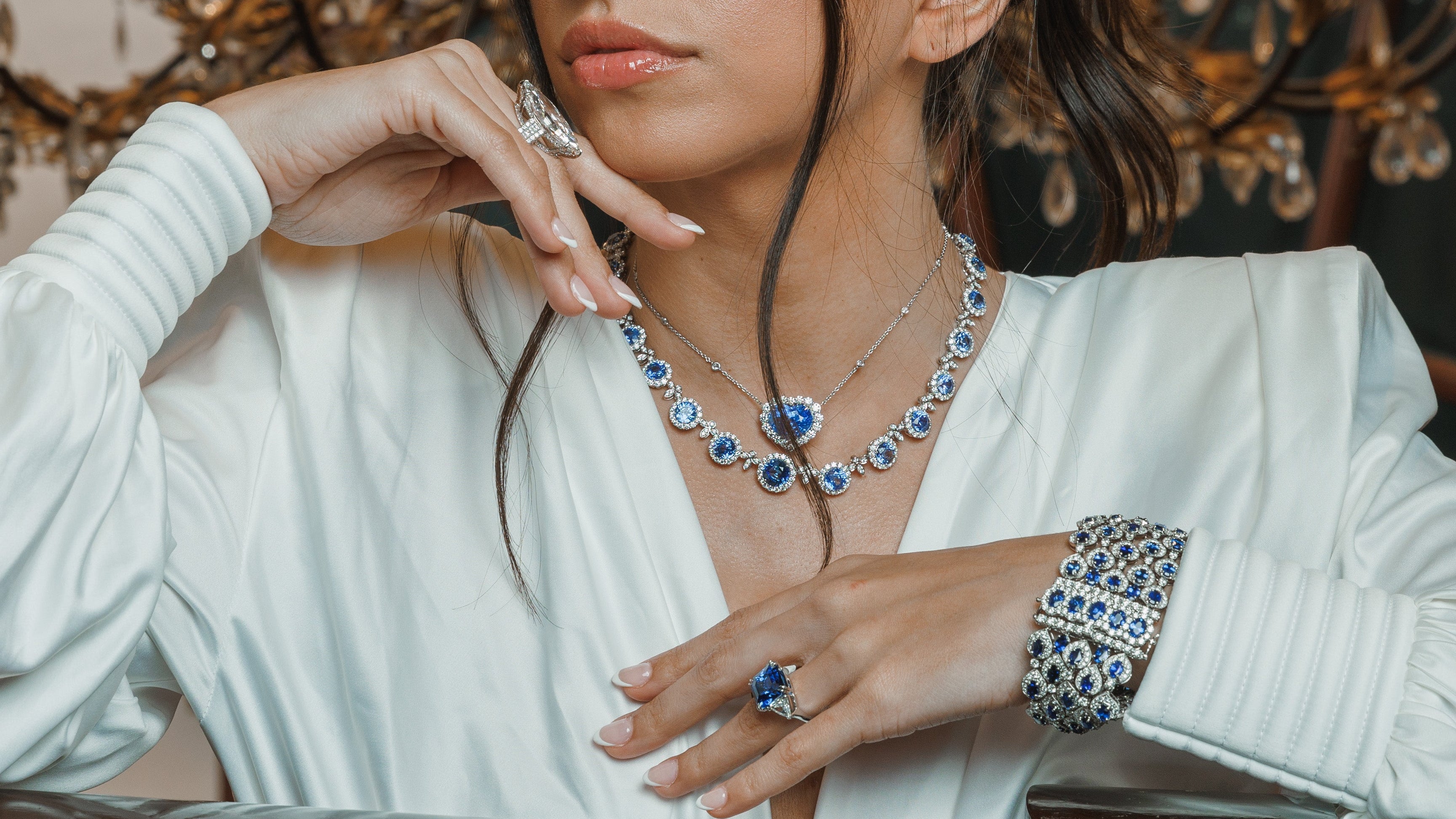 A woman in a white blouse leans on a wooden table, wearing a blue and silver necklace and matching rings, with a large lit chandelier in the background.