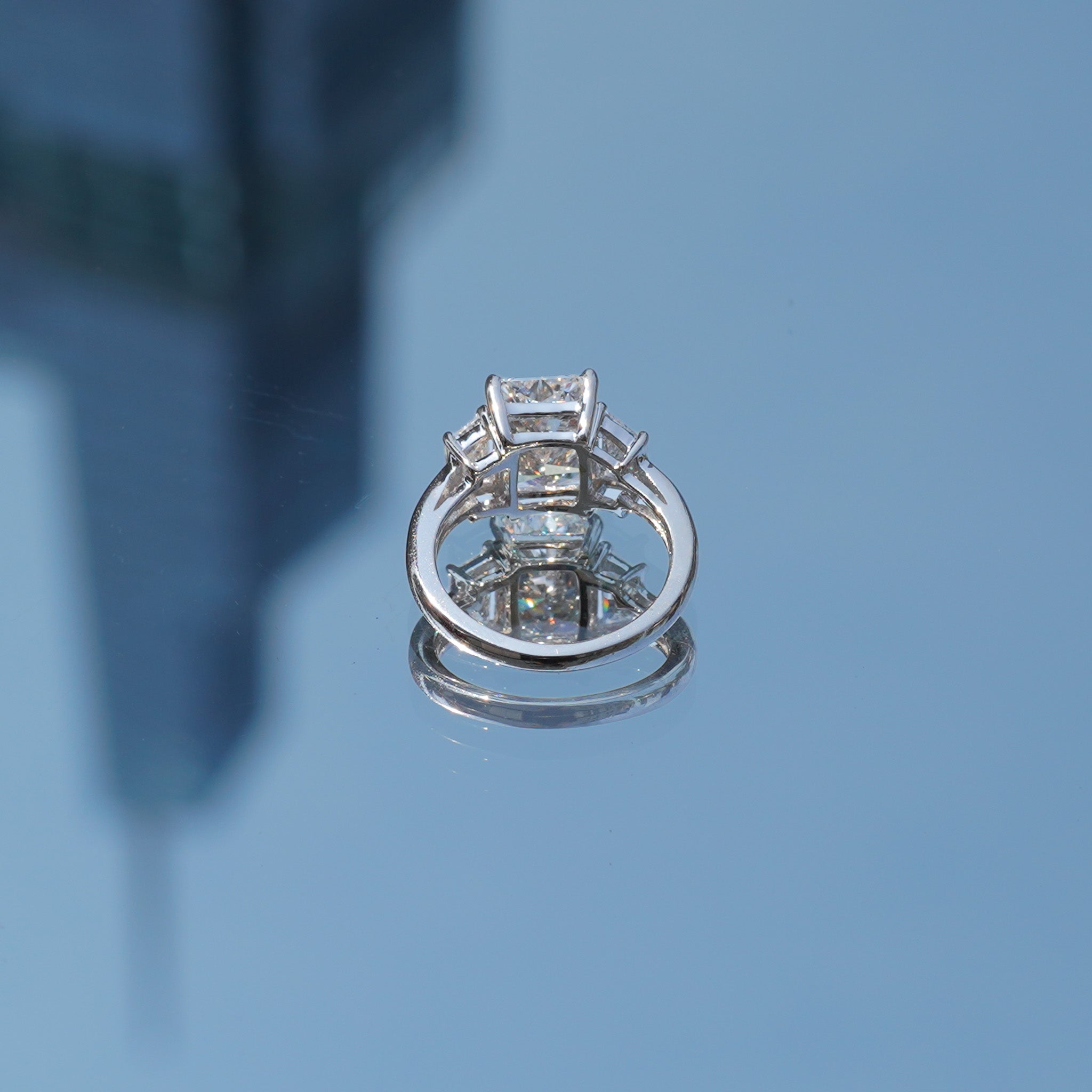 A silver-toned ring with a large rectangular clear gemstone and smaller accent stones, photographed on a reflective blue surface with a blurred background.