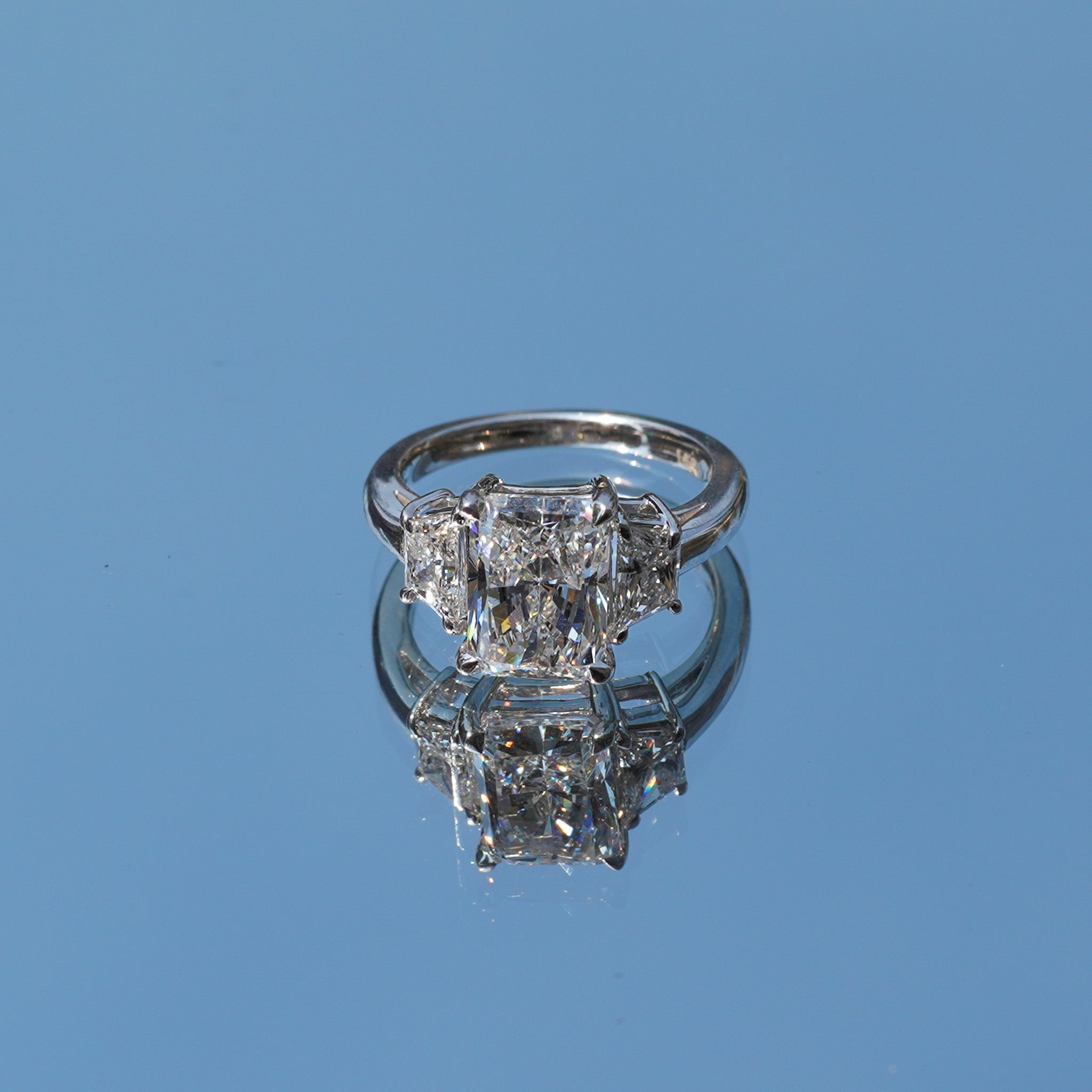 A silver three-stone diamond ring with a central round brilliant-cut diamond flanked by two smaller baguette-cut diamonds, displayed on a reflective blue surface.