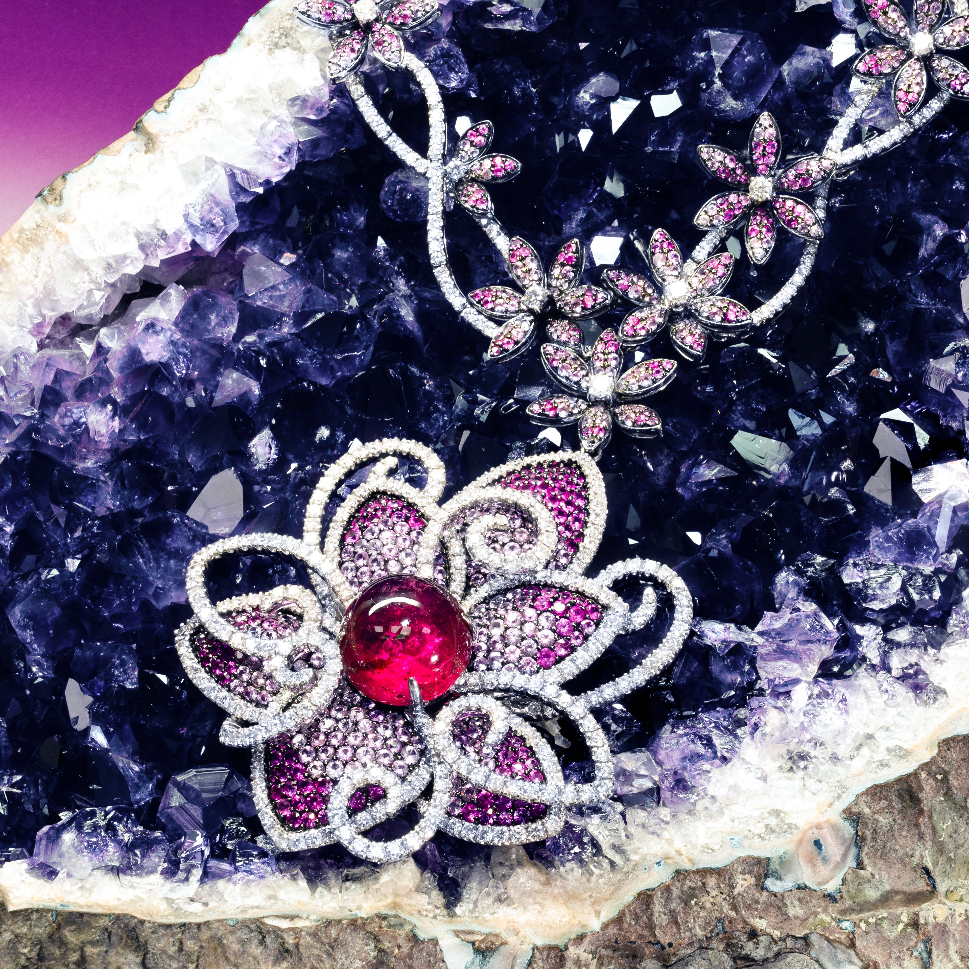 A close-up of a floral brooch with silver-toned filigree and pink gemstone petals, set with a central red gemstone, displayed on a purple crystal geode.