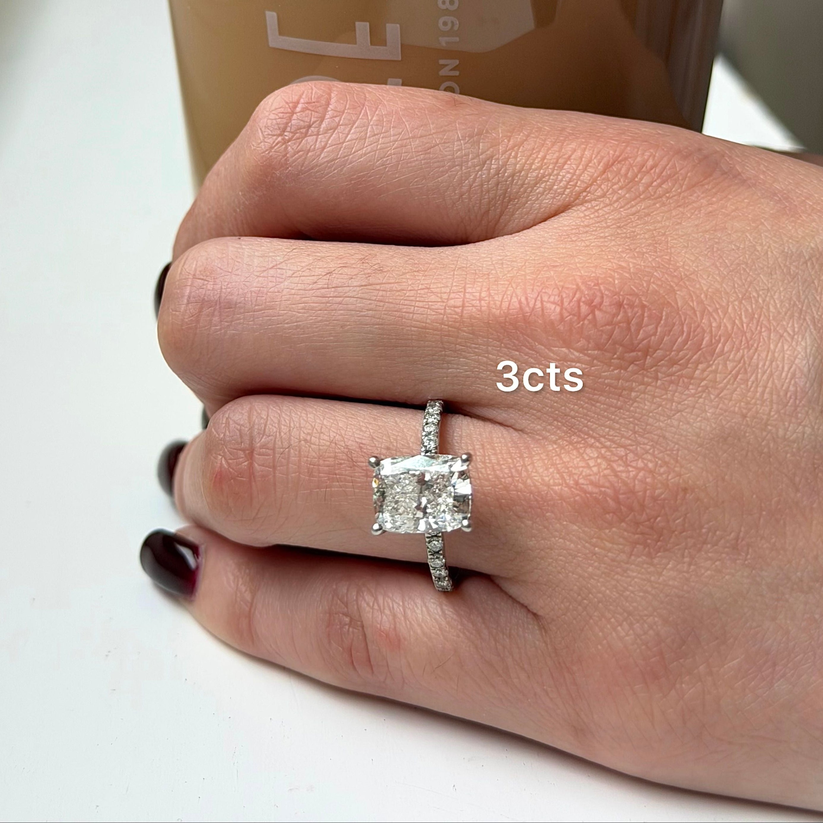 Hand wearing a diamond ring with '3 cts' next to a cup of coffee