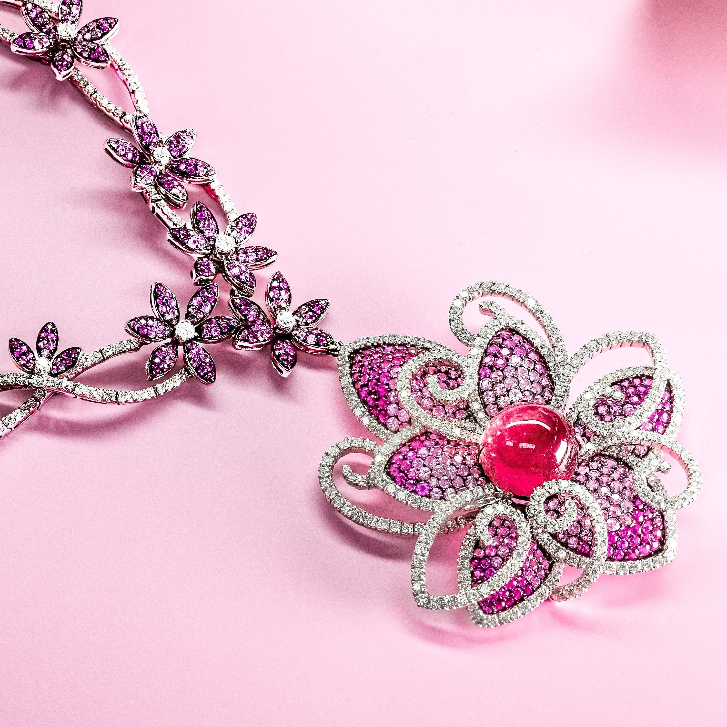 A floral filigree necklace with a large flower pendant, featuring pink gemstones, and a central red cabochon, displayed on a soft pink background with decorative silver leaves.