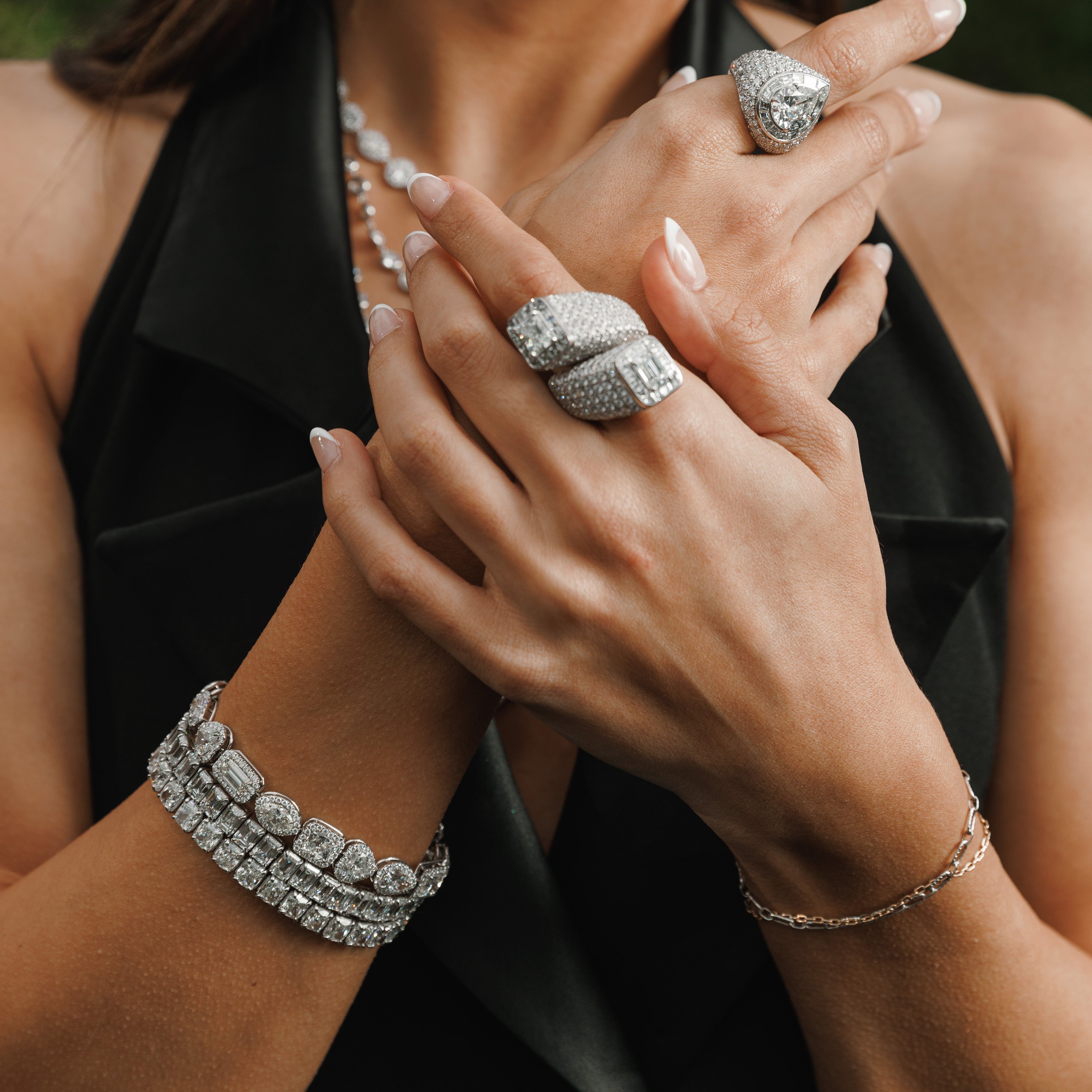 A close-up lifestyle shot of a woman wearing multiple silver-toned jewelry pieces, including a large ring, hoop earring, bracelet, and necklace, with a textured black sleeveless top.