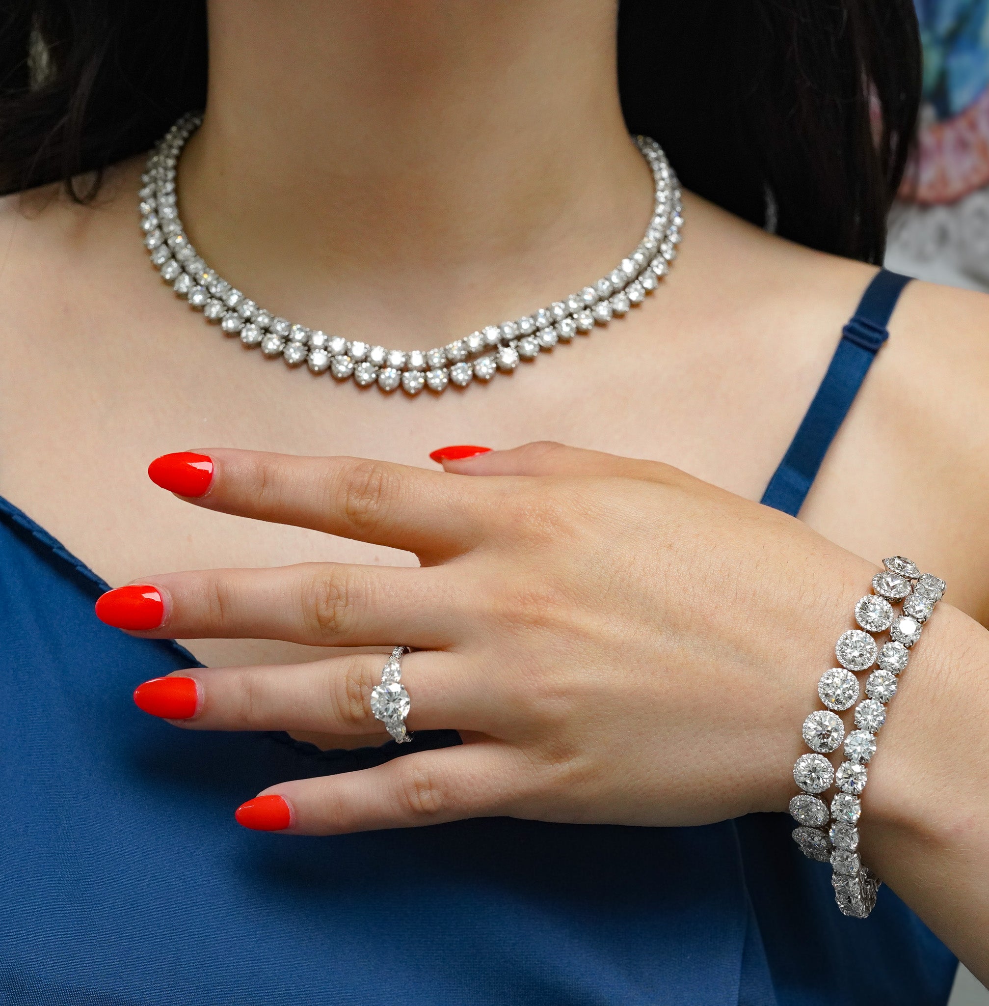 Close-up of a person wearing a diamond necklace, ring, and bracelet with red nail polish.