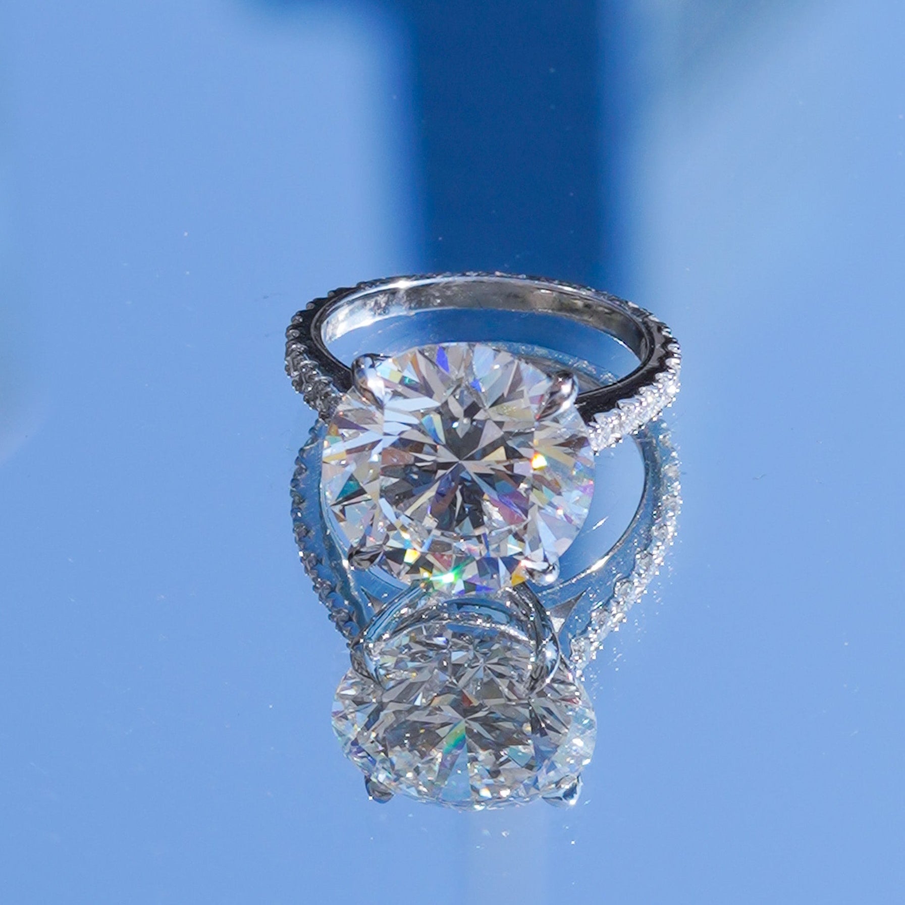 A ring with a large round brilliant-cut clear center stone and a halo of small clear stones, photographed on a reflective blue surface with a mirrored reflection.