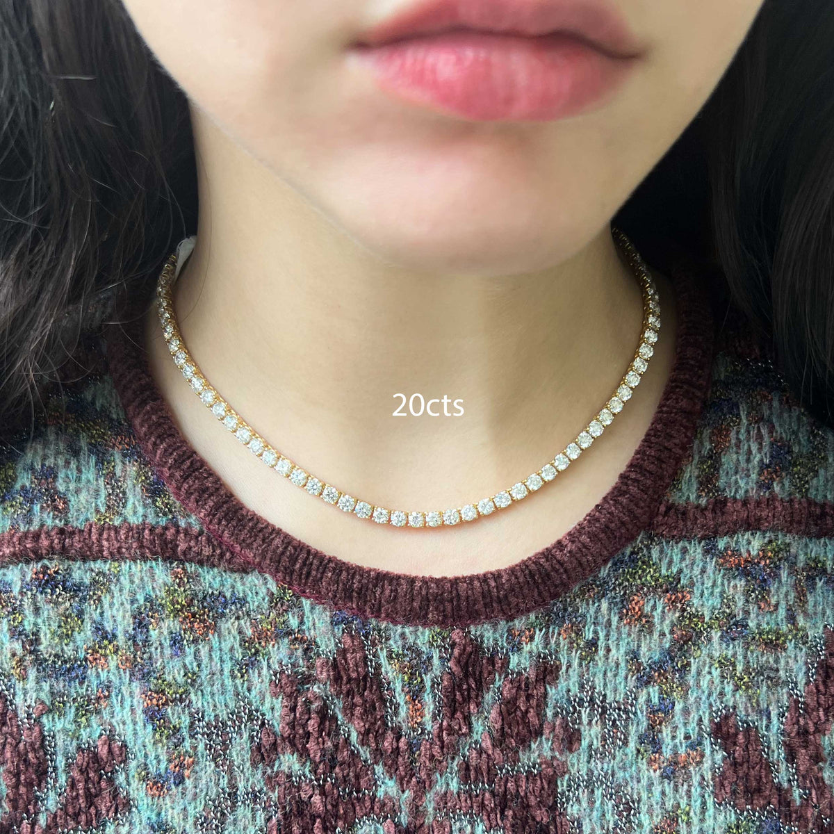 A close-up of a person's neck and lower face wearing a delicate tennis-style necklace with clear stones set in a yellow gold-tone band. The person is wearing a textured knit sweater with a burgundy and blue pattern.