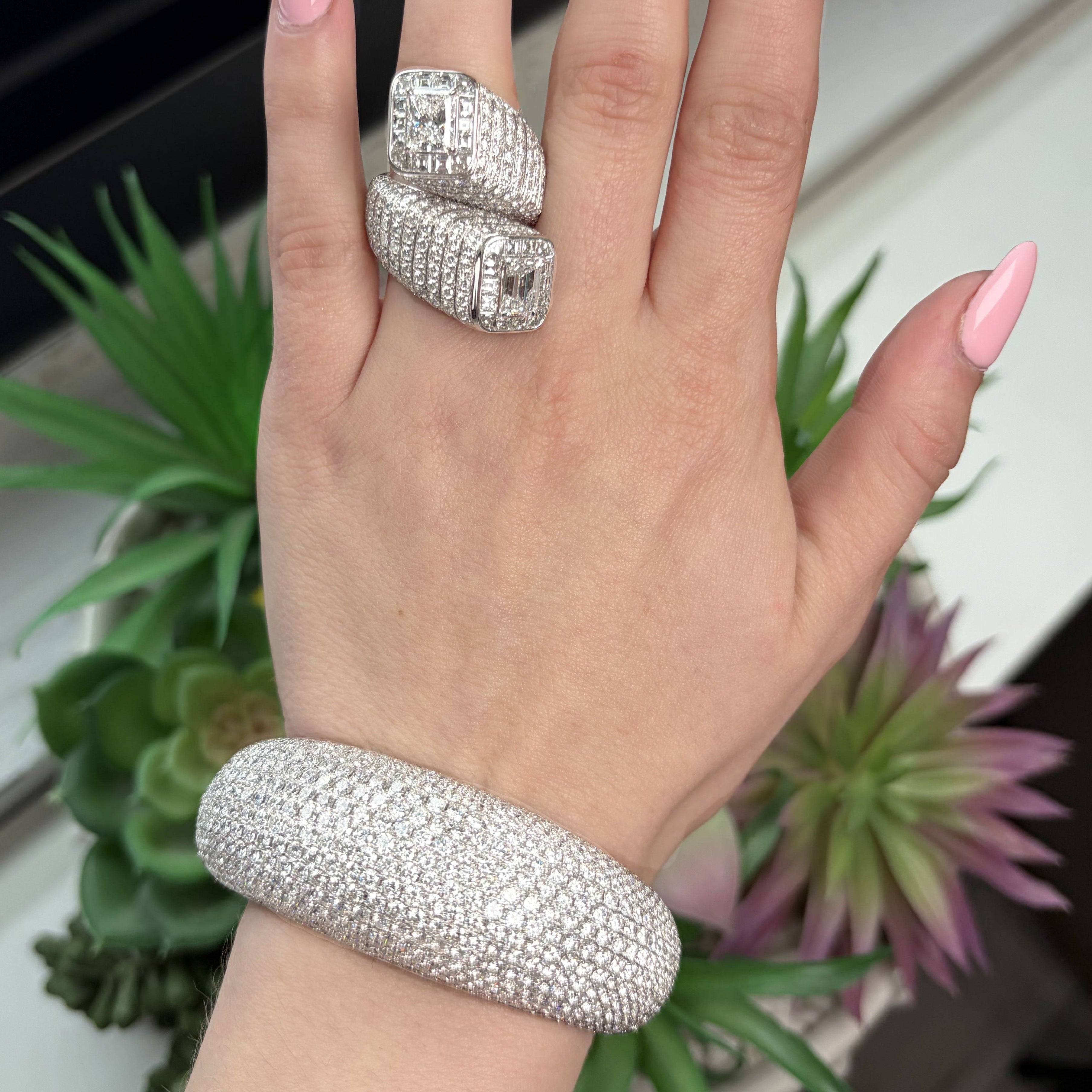 Hand wearing a silver ring and bracelet with pink nail polish, against a neutral background.