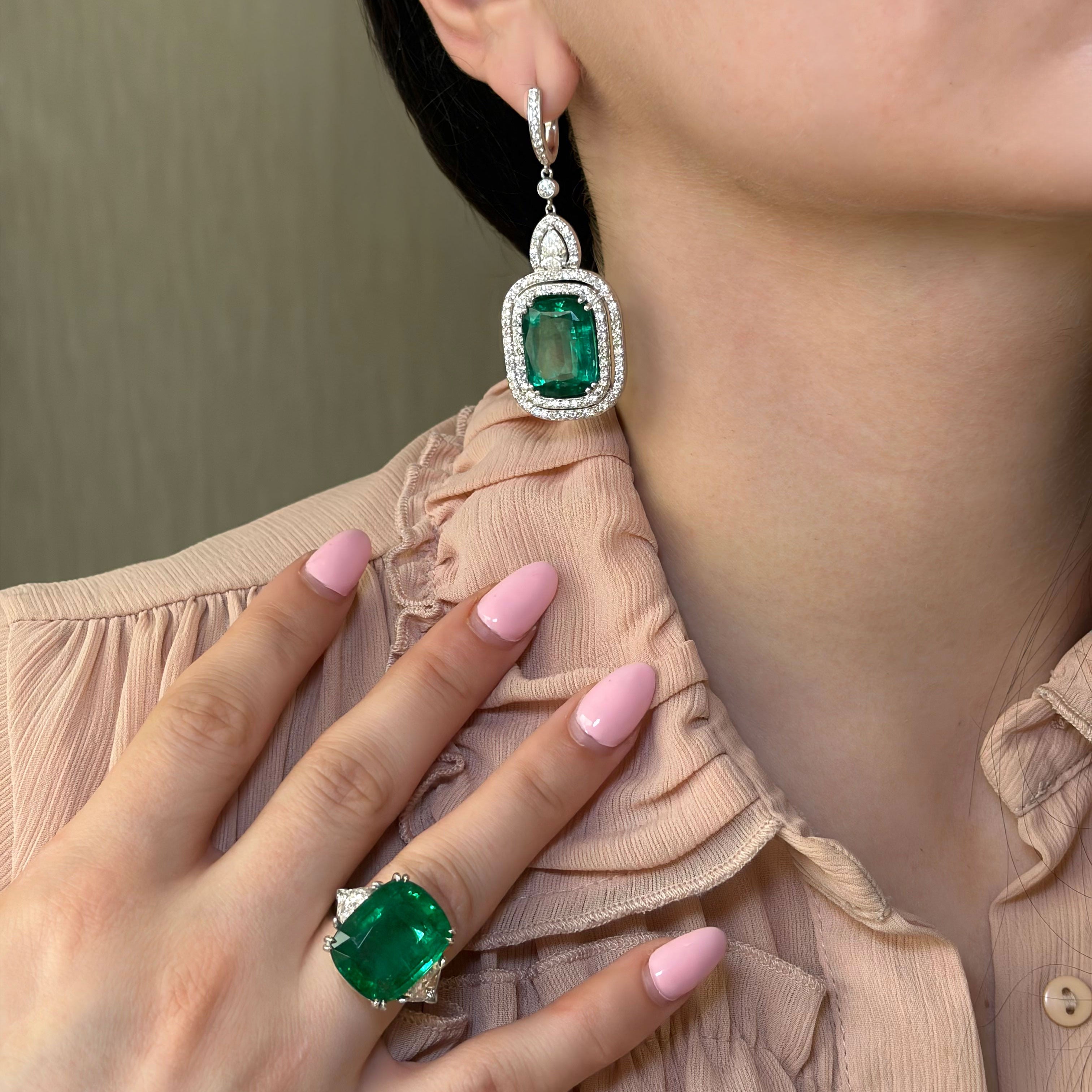 Close-up of a person wearing emerald green earrings and a ring, with a neutral background.