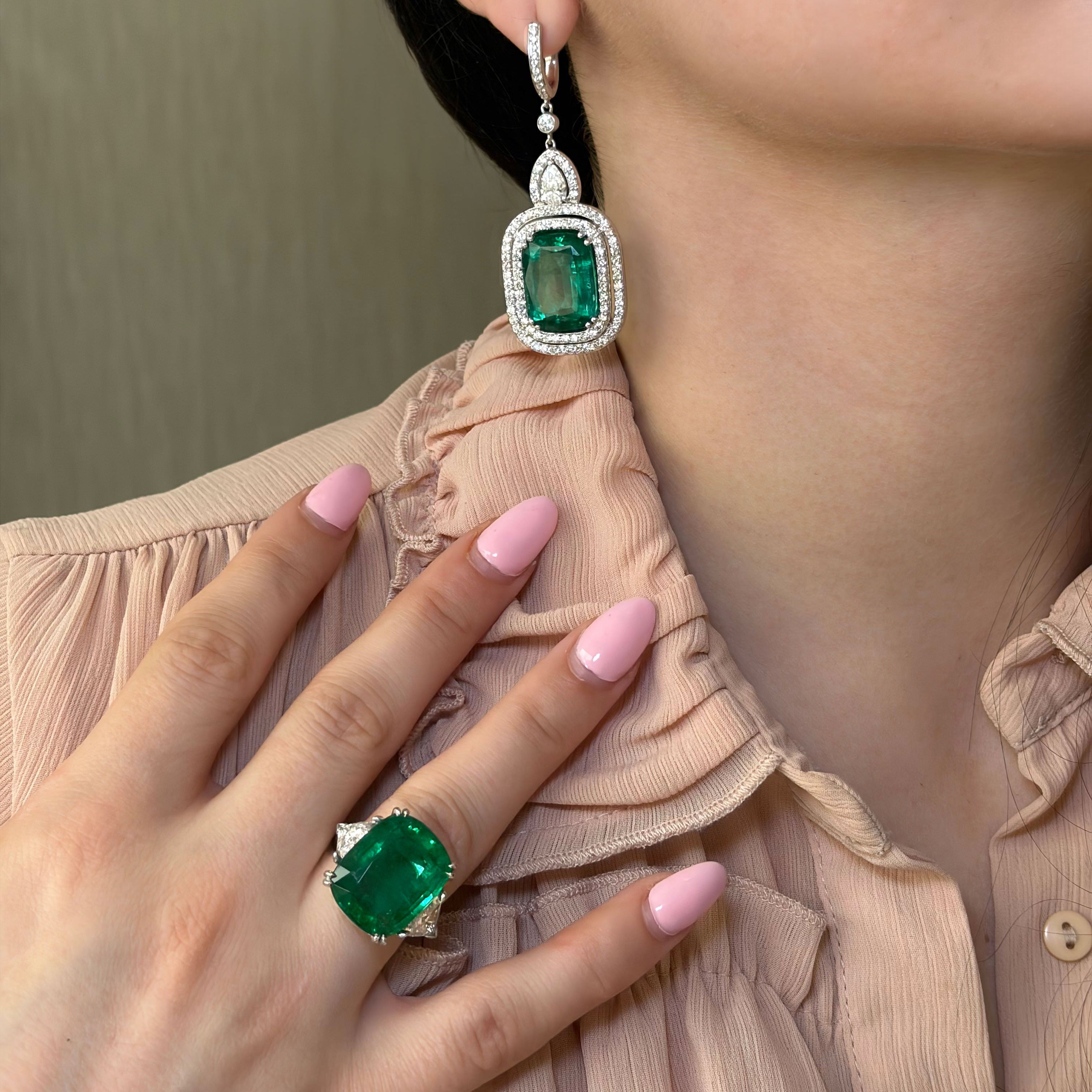 Close-up of a person wearing emerald green earrings and a ring, with a neutral background.